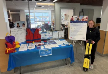 YMCA staff member standing beside a National Drowning Prevention Week information table displaying lifejackets, water safety resources, swimming lesson information, and aquatic certification materials in a YMCA lobby.