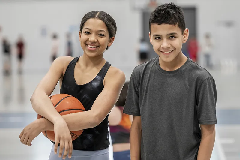 [image] A teen boy and girl stand in the gym. The girl holds a basketball.