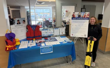 YMCA staff member standing beside a National Drowning Prevention Week information table displaying lifejackets, water safety resources, swimming lesson information, and aquatic certification materials in a YMCA lobby.